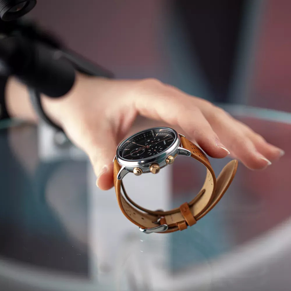 Mão posicionando relógio masculino com pulseira marrom de couro sobre superfície de vidro